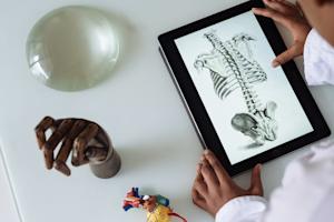 A person using a tablet to remotely access a school lab computer. The tablet screen shows the skeletal structure of the human body.