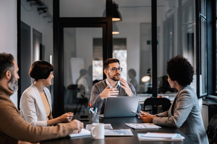 Four professionals sit around a table in a modern office, engaged in discussion. One man speaks while the others listen attentively. A laptop, documents, and coffee cups are on the table.