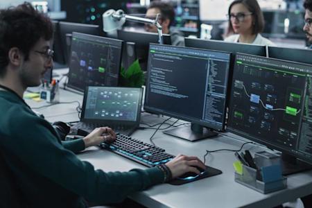 A person works at a desk with multiple computer monitors displaying code and technical diagrams, while three colleagues sit at nearby workstations in a modern office setting.