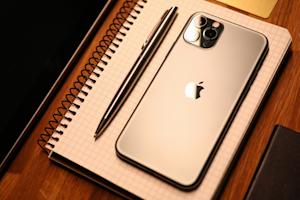 A silver iPhone on a spiral notebook with a pen, placed on a wooden desk.