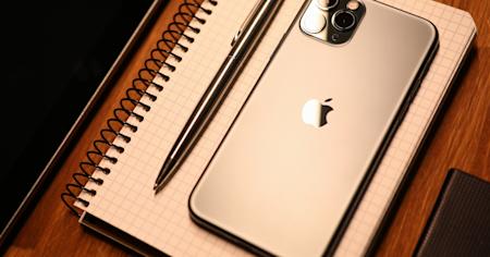 A silver iPhone on a spiral notebook with a pen, placed on a wooden desk.