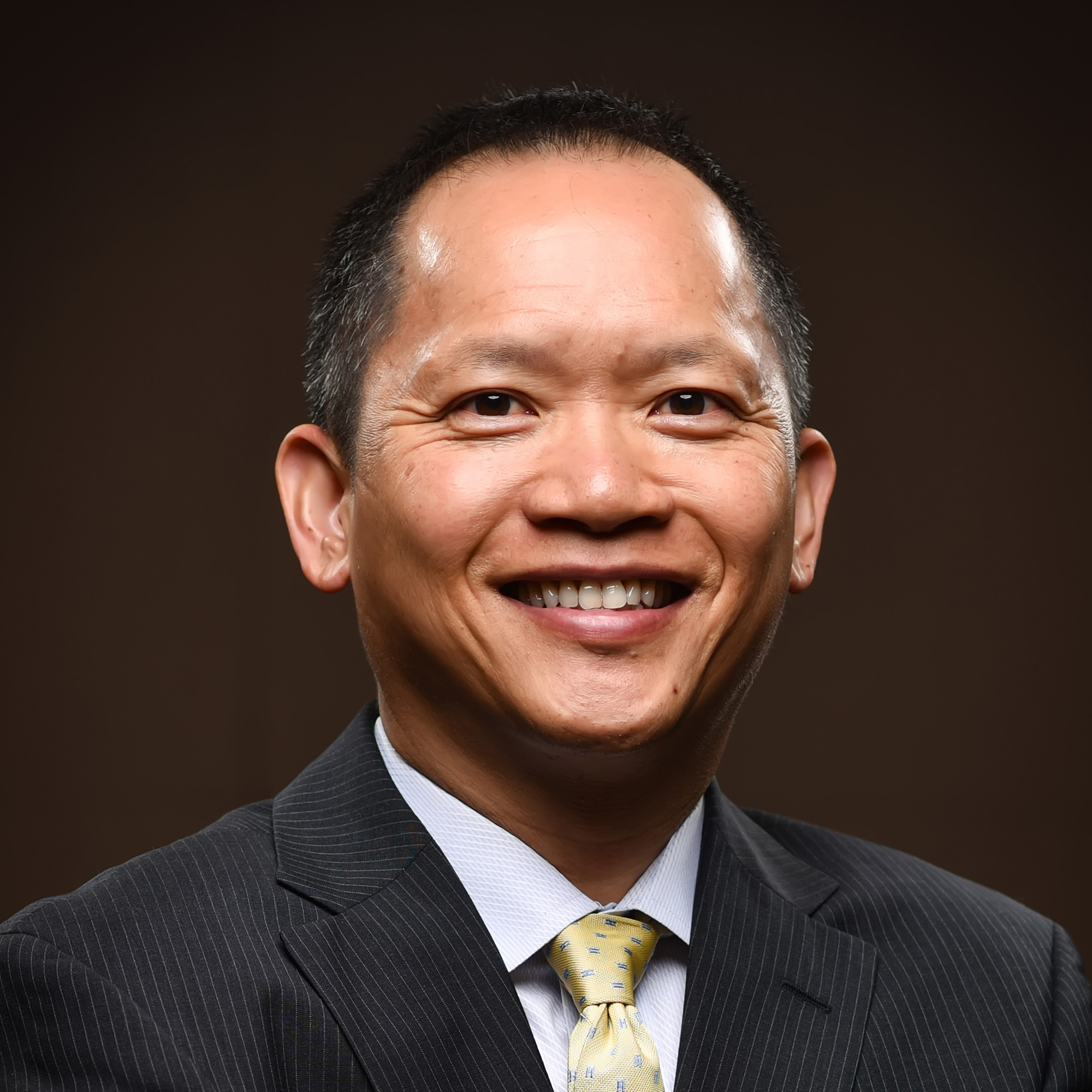 A smiling man in a dark pinstripe suit, light blue shirt, and yellow patterned tie poses against a plain dark background.