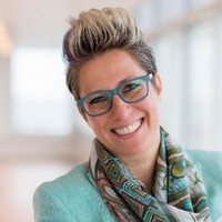 Smiling person with short, spiked hair and blue glasses, wearing a turquoise jacket and a patterned scarf, standing in a bright, blurred indoor setting.