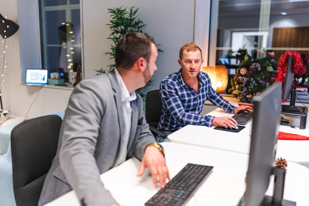 Two employees talking to each other and working on their computers.