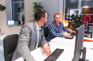 Two employees talking to each other and working on their computers.