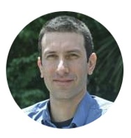 A man with short dark hair and light skin wearing a blue collared shirt is outdoors with greenery in the background.