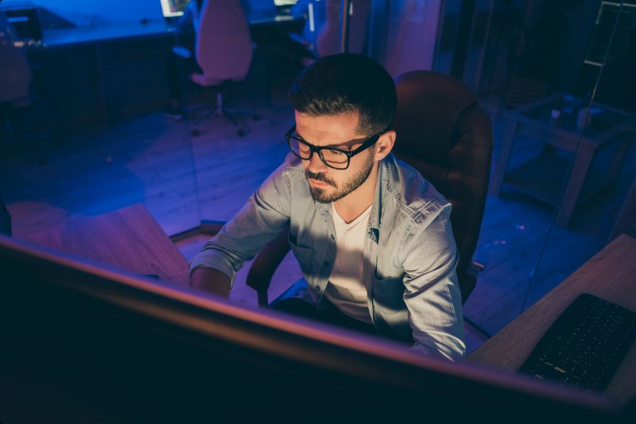 A man with dark hair and glasses sits at a desk, working on a computer in a dimly lit room with a blue glow. He appears focused, wearing a light shirt, with computer monitors and chairs visible in the background.