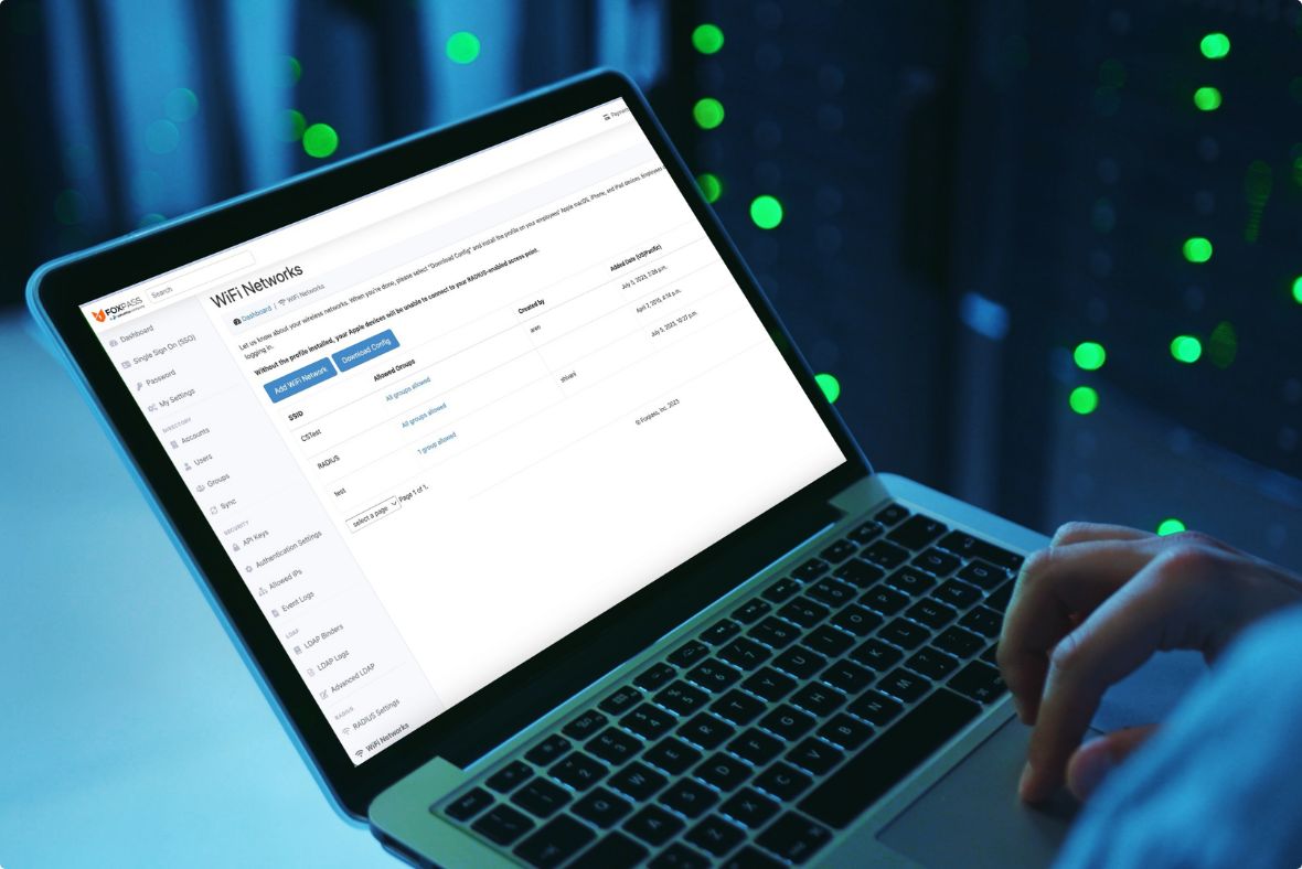 A person uses a laptop in a dim server-room environment; the screen shows a web-based admin dashboard with a list of user accounts and settings, illuminated by green indicator lights from nearby server racks.