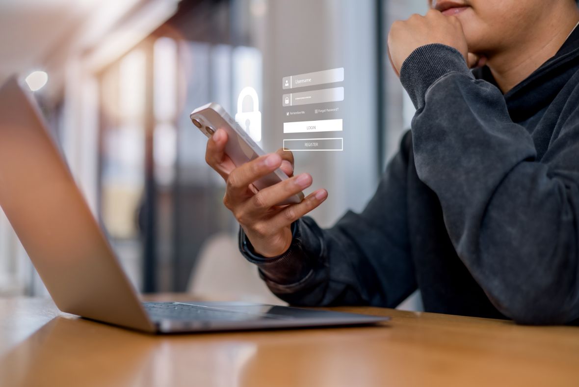 A person sits at a desk with a laptop, holding a smartphone. A transparent login form is projected in the air, suggesting a digital or online login process. The person appears thoughtful and focused.