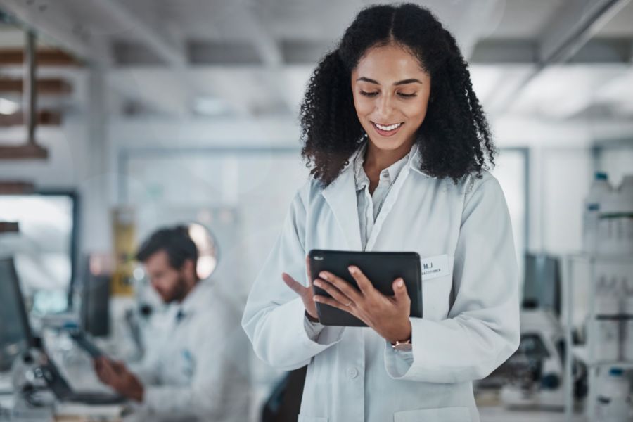 A woman in a white lab coat smiles while using a tablet in a laboratory. Another person in a lab coat works in the background. The lab environment includes equipment and shelves.