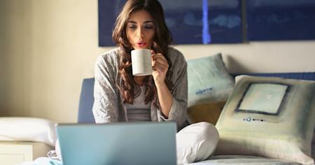 A woman working from home on her laptop while sitting on a bed and drinking coffee.