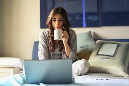 A woman working from home on her laptop while sitting on a bed and drinking coffee.