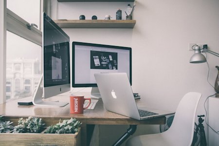 A Mac laptop on a desk in front of two other Apple monitors.