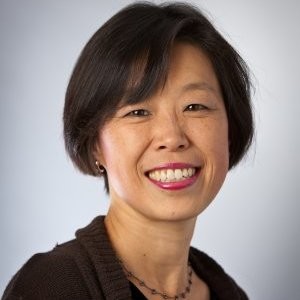 A woman with short black hair and a warm smile, wearing a dark sweater and a necklace, is pictured against a plain, light background.