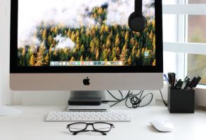 A Mac desktop computer on a desk with a keyboard, mouse, and a pair of glasses.