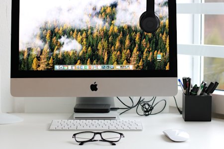 A Mac desktop computer on a desk with a keyboard, mouse, and a pair of glasses.