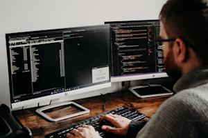 A person wearing glasses types on a keyboard while looking at two computer monitors displaying code and file directories in a dimly lit workspace.