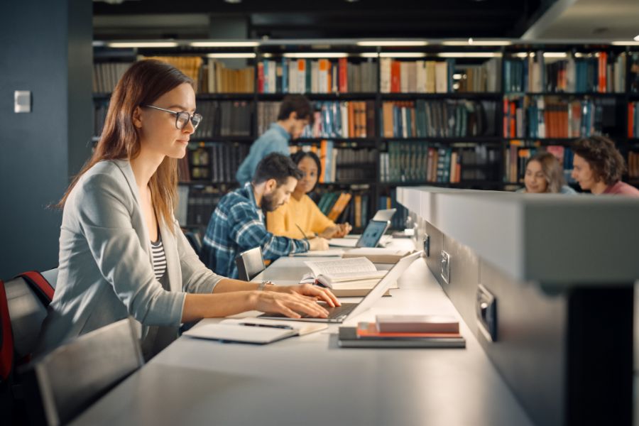 A woman wearing glasses studies at a desk in a library, surrounded by books and a laptop. In the background, several other people are also reading and working at tables lined with bookshelves.