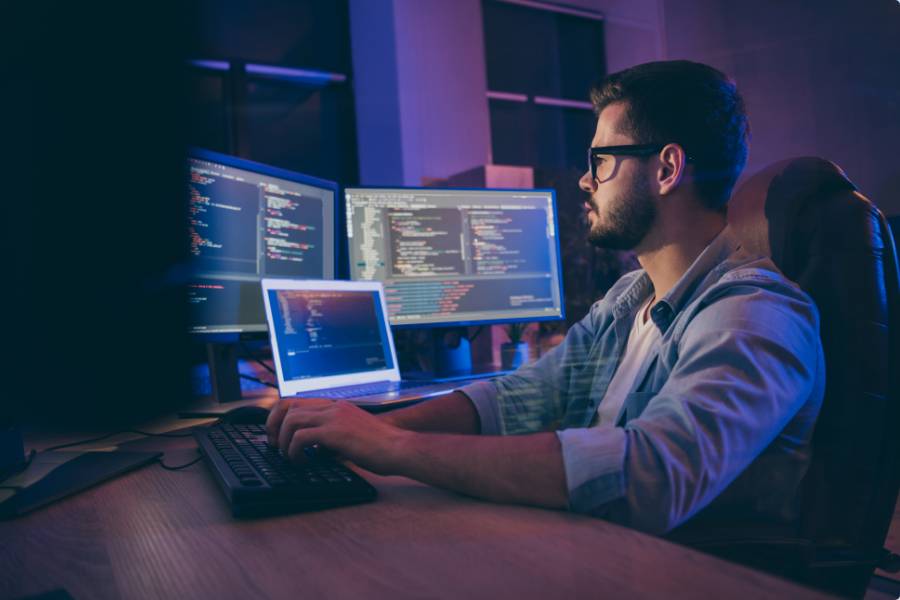 A person sits at a desk in a dark, neon-lit workspace, typing on a keyboard while multiple monitors display lines of code and development tools.
