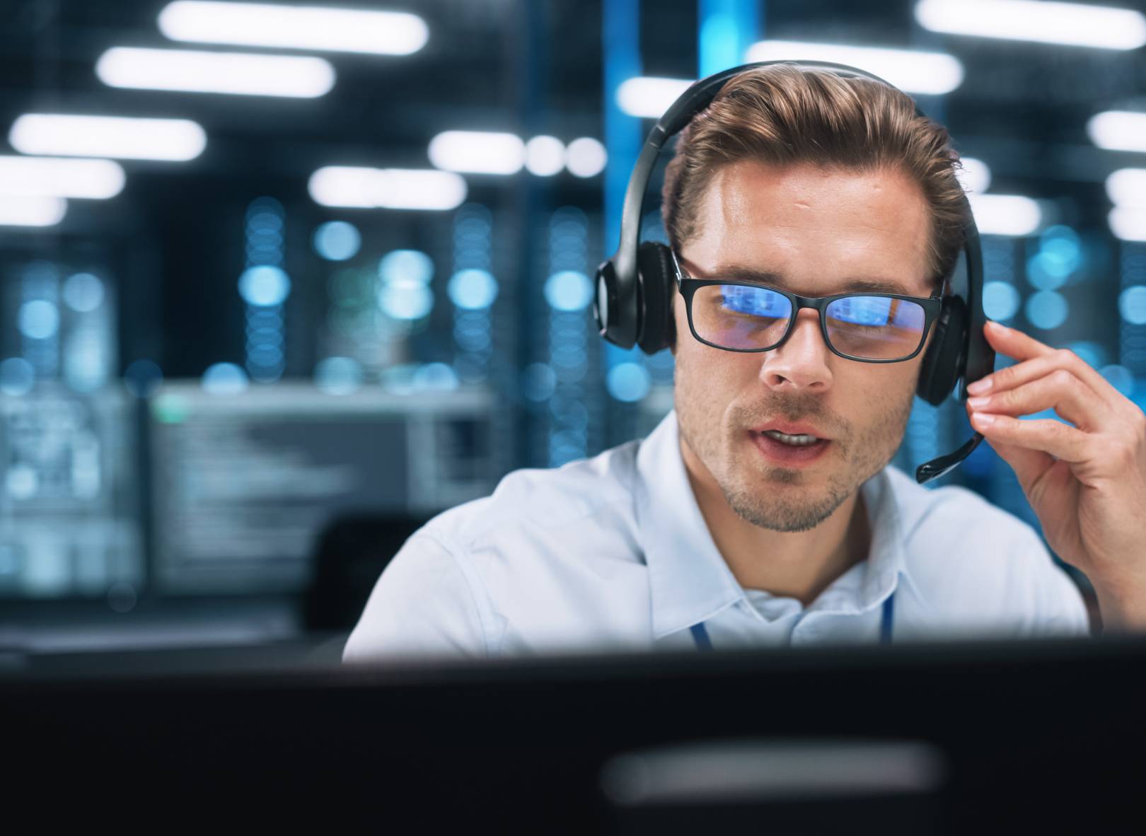 A man wearing glasses and a headset works at a computer in a modern office with multiple screens and blue LED lighting in the background.