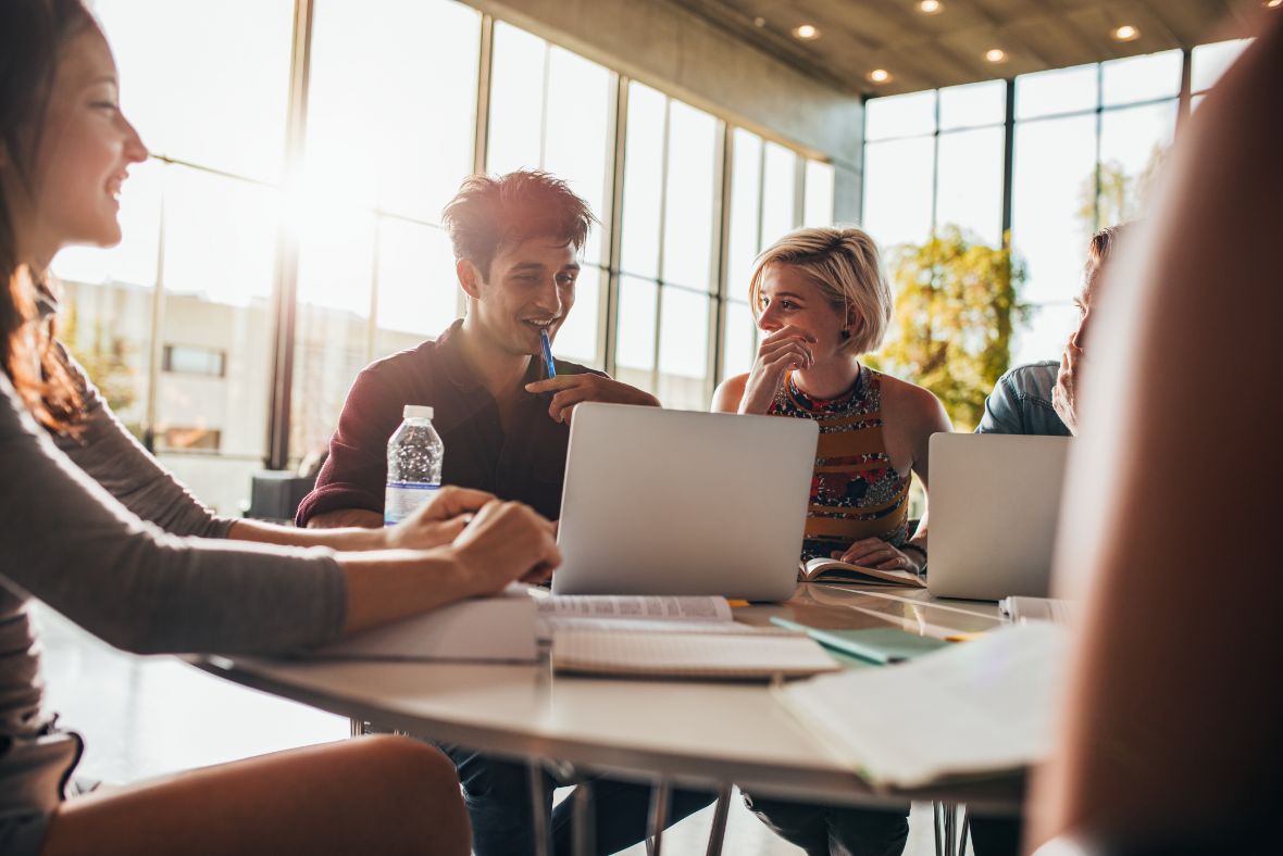 Four young adults sit around a table with laptops, notebooks, and a water bottle, smiling and laughing while working together in a bright, modern office space with large windows.