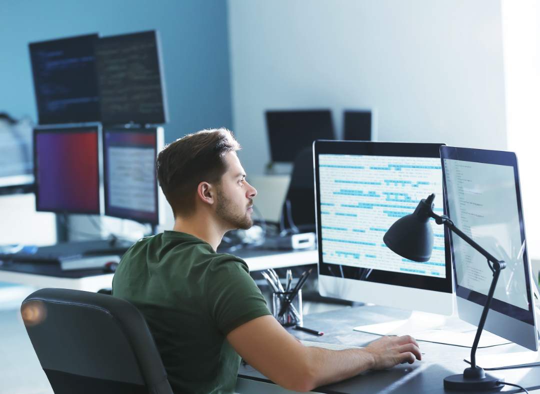 A man sits at a desk in an office, working on a computer with multiple monitors displaying code. A desk lamp, pens, and other office supplies are nearby. Sunlight streams in from a large window.