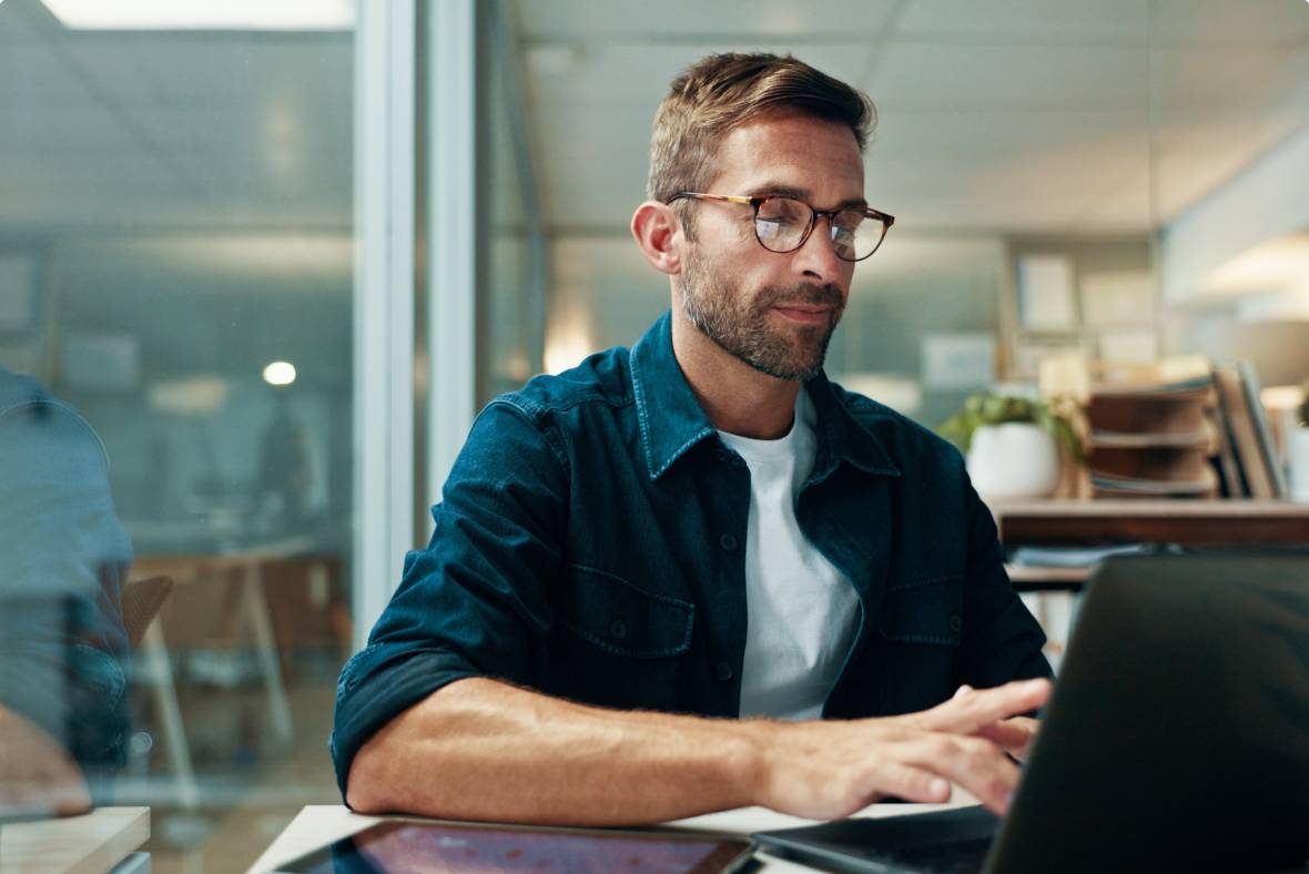 A man wearing glasses and a denim shirt sits at a desk using a laptop in a modern office. There are shelves, plants, and documents in the background, and a tablet is on the desk in front of him.