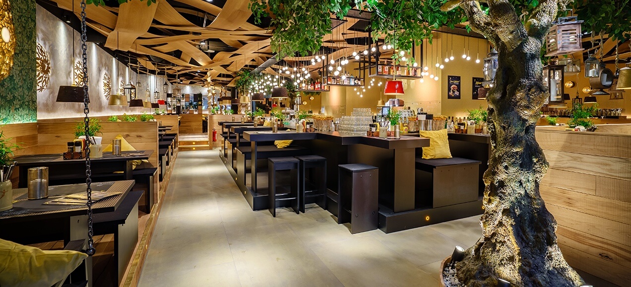A modern restaurant interior with wooden tables, black chairs, hanging lights, indoor plants, and a large decorative tree, creating a warm and inviting atmosphere.