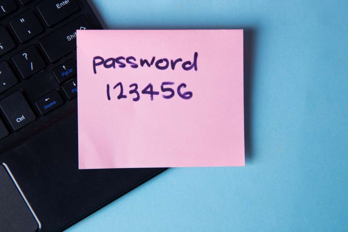 A pink sticky note with password 123456 written on it is placed on a black laptop keyboard, against a light blue background.