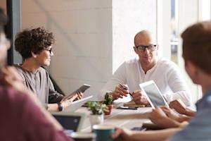 A group of coworkers talking at a conference table while working on their devices.