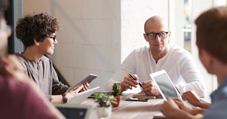 A group of coworkers talking at a conference table while working on their devices.