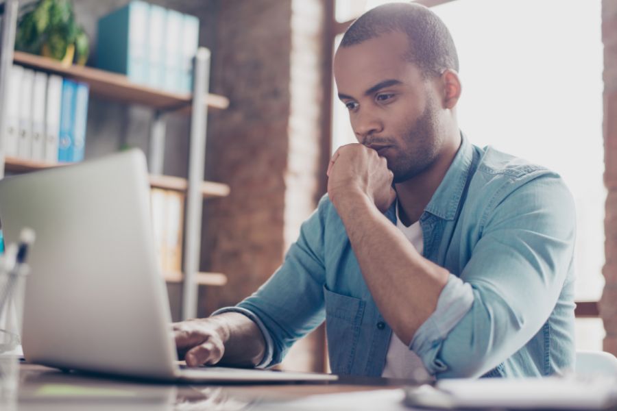 A man sits at a desk, looking thoughtfully at a laptop screen with his hand resting on his chin. He is wearing a light blue shirt and appears focused, with bookshelves and office supplies in the background.