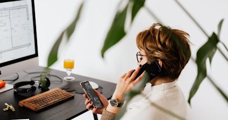 A worker on the phone in front of a desktop computer looking at a different mobile device in their hand while receiving remote support.
