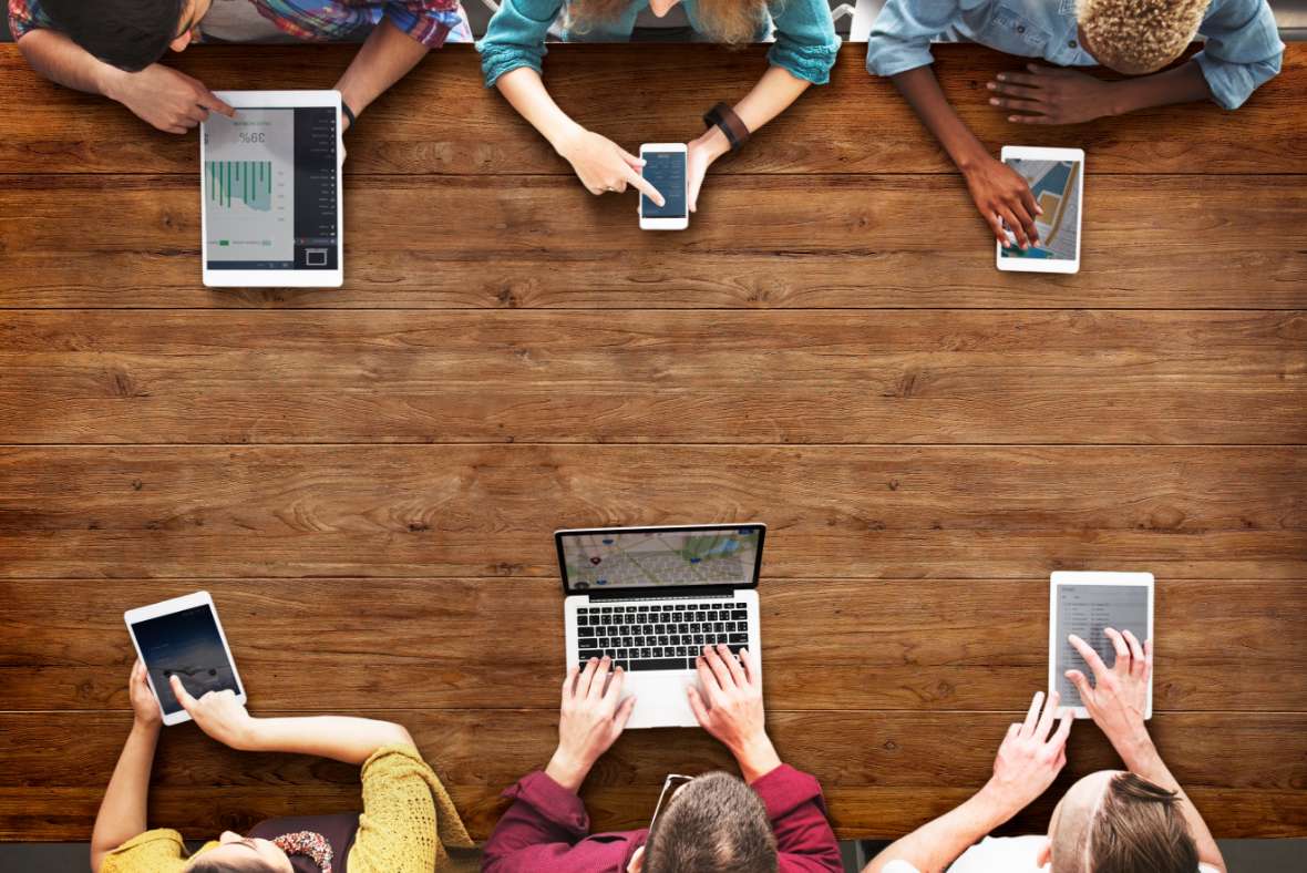Six people sit around a wooden table using various devices—laptops, tablets, and smartphones—seen from above, with their hands interacting with the screens. The center of the table is empty.