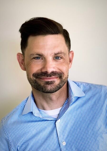 A man with short dark hair and a beard smiles at the camera. He is wearing a light blue and white checkered button-up shirt over a white t-shirt, and is posed against a plain light-colored background.