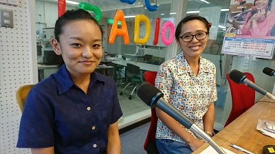 Two women sit in a radio studio in front of microphones, smiling at the camera. Colorful balloon letters spelling RADIO are visible in the background. The studio has glass walls and audio equipment.