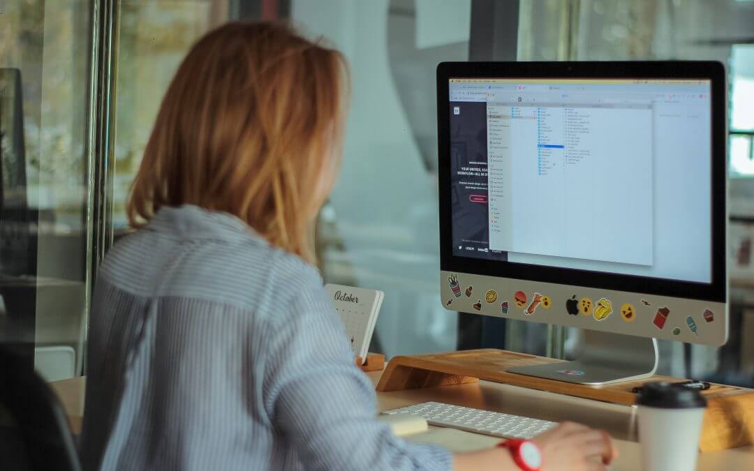 A person with light brown hair sits at a desk using a desktop computer with a screen displaying code or text files. A coffee cup, notebook, and other items are on the desk in a modern office setting.