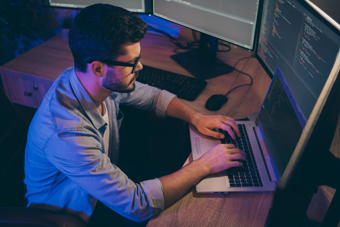 A man with glasses types on a laptop at a desk, surrounded by multiple monitors displaying code in a dimly lit room.