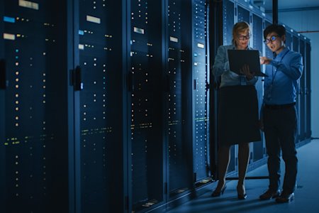 Two IT professionals reviewing data on a laptop in a server room filled with tall racks of network equipment.