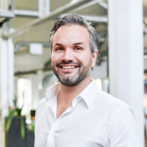 A smiling man with short, gray-streaked hair and a trimmed beard wears a white button-up shirt. He stands indoors in a modern, bright office space with blurred backgrounds.