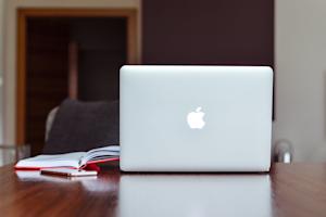 A Mac laptop on a desk next to a notebook.