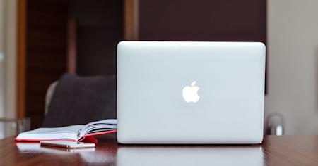 A Mac laptop on a desk next to a notebook.