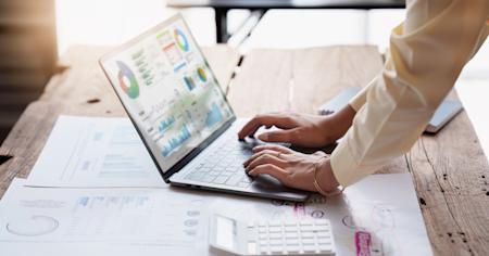 Person using a laptop with workflow management charts on a desk.