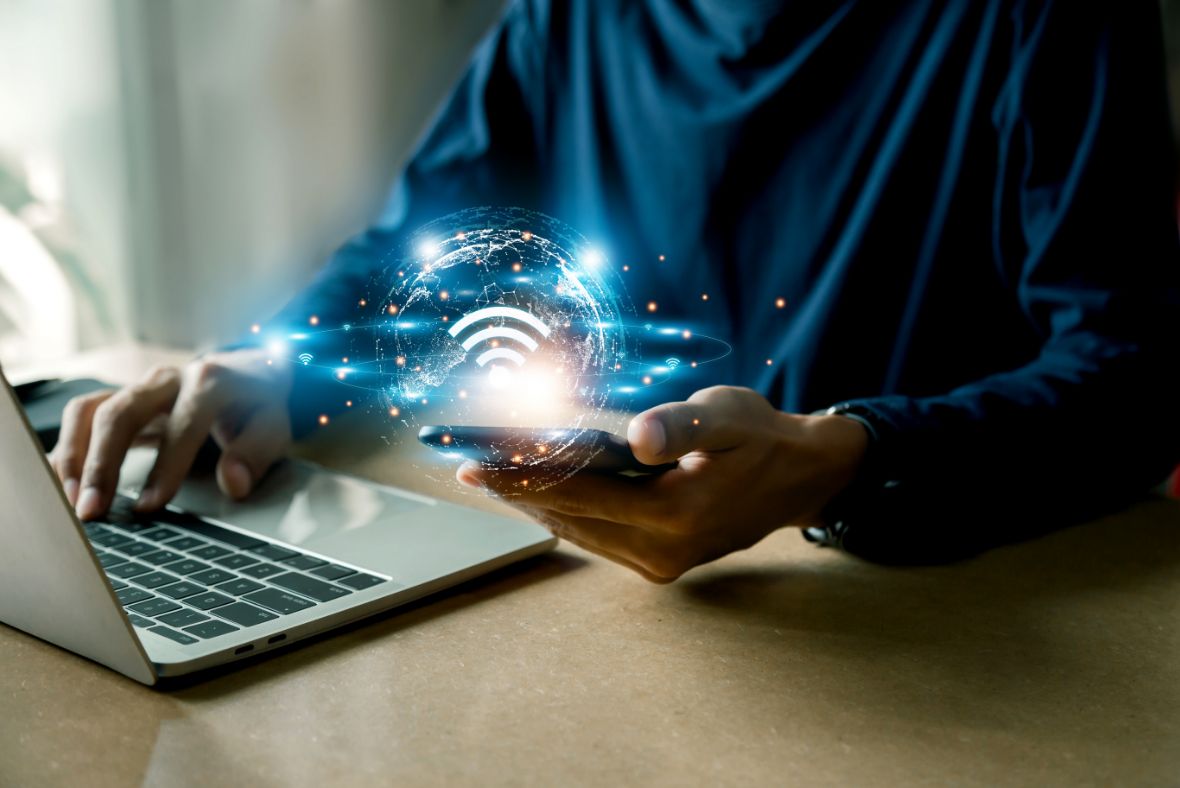 A person using a laptop and holding a smartphone, with a glowing holographic globe and Wi-Fi symbol appearing above the phone, representing wireless connectivity and internet technology.
