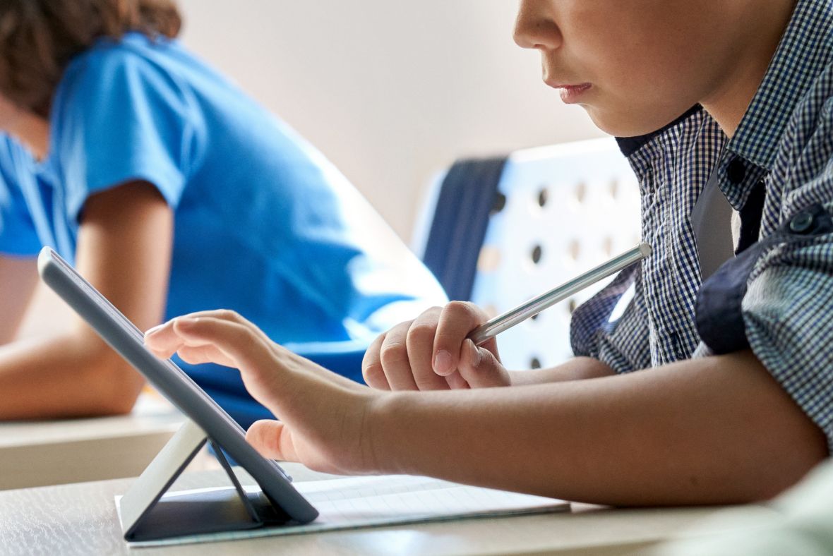 A child using a tablet with a stylus in a classroom, focusing on the screen. Another student sits nearby, slightly out of focus.