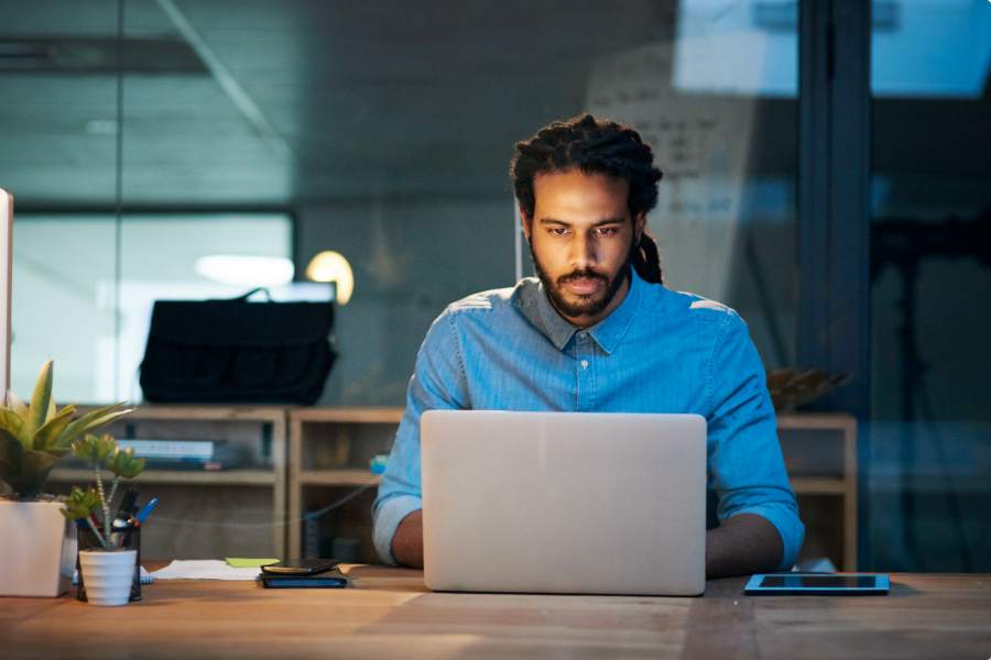 A man with braided hair sits at a desk in a modern office, focused on his laptop. There is a smartphone, a tablet, and a plant on the wooden desk, and shelves in the background.