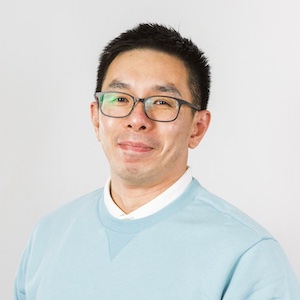A man with short black hair and glasses is wearing a light blue sweater over a white collared shirt. He is smiling softly and standing in front of a plain, light gray background.