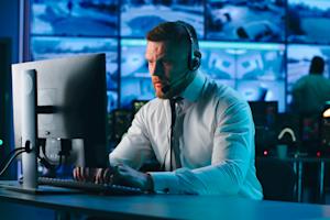 IT professional in a control room using a computer and headset, managing hybrid IT infrastructure with multiple screens displaying data in the background.