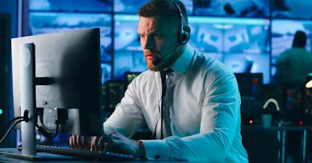 IT professional in a control room using a computer and headset, managing hybrid IT infrastructure with multiple screens displaying data in the background.