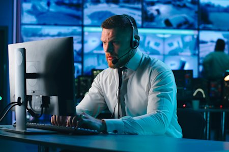 IT professional in a control room using a computer and headset, managing hybrid IT infrastructure with multiple screens displaying data in the background.